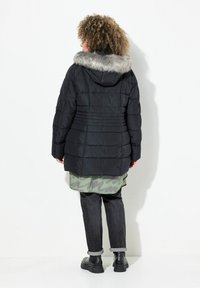 Chaqueta negra acolchada con capucha de felpa gris desmontable, textura acolchada y cintura ajustada; llevada sobre una camisa verde con estampado y jeans oscuros.
