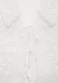 Blouse blanche avec broderie florale, décolleté en V et fermeture à nouer. La texture est douce avec un tissu délicat et léger.