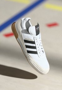 Białe sneakersy Adidas Mundial Goal z czarnymi paskami i gumową podeszwą, zawieszone nad jasną podłogą z kolorowymi liniami.