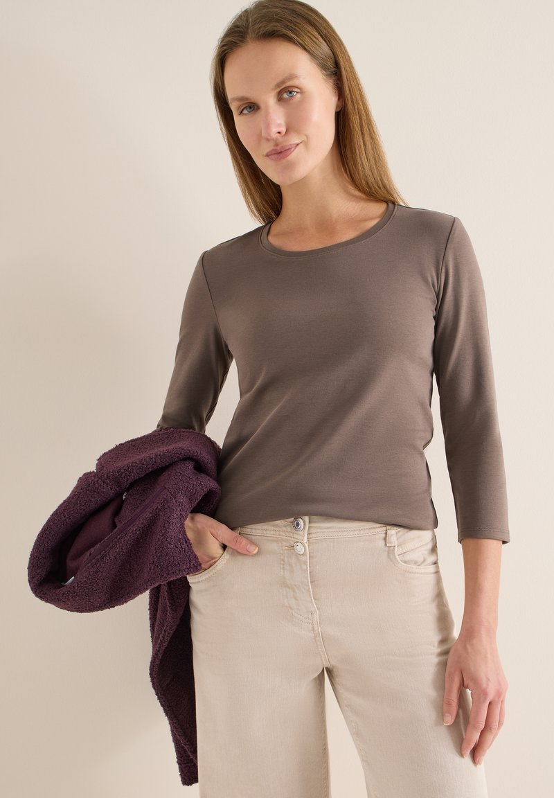 Haut marron à manches longues avec un col rond, associé à un jean clair à coupe décontractée. Une veste violette texturée est posée sur le bras.