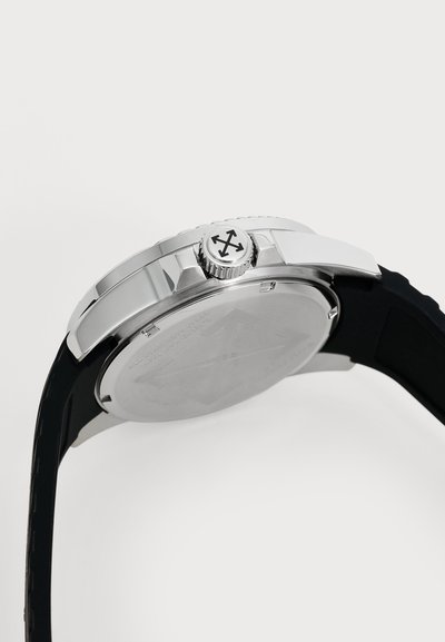 Gros plan sur le boîtier argenté d’une montre-bracelet et son bracelet en caoutchouc noir, vus de côté, avec la couronne et le logo à quatre flèches visibles.