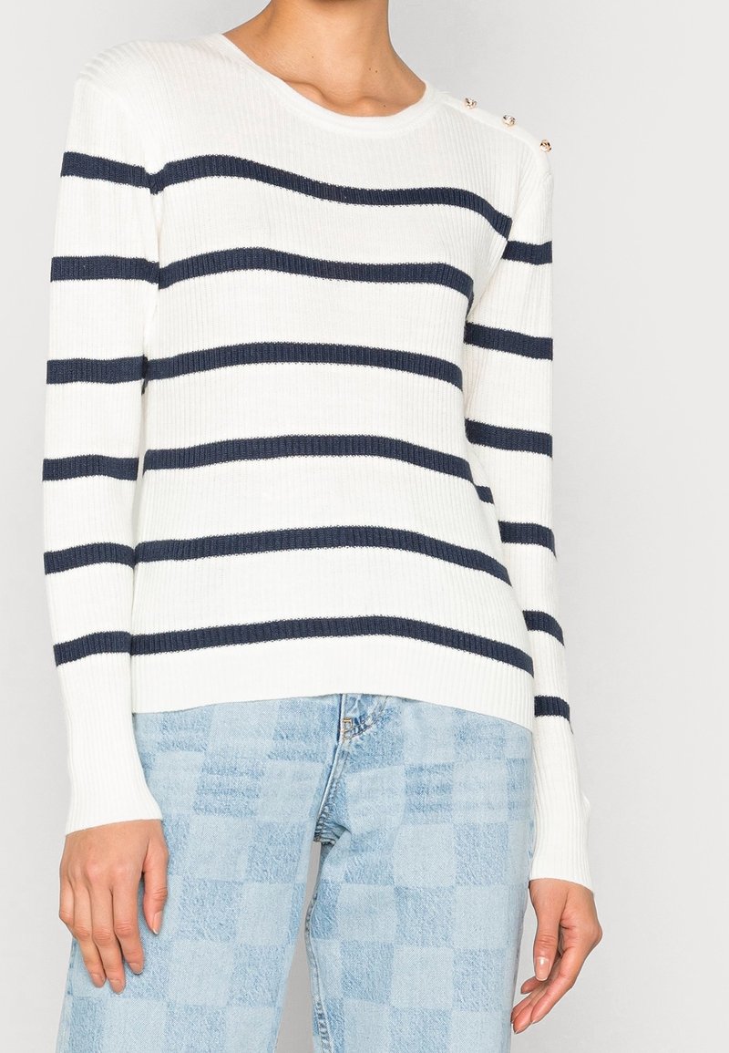Femme portant un pull blanc côtelé avec des rayures horizontales noires, des boutons dorés sur une épaule, et un jean bleu clair à carreaux.