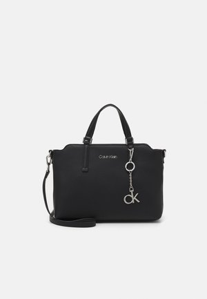 Sac à main Calvin Klein noir avec doubles poignées, bandoulière amovible et pendentif logo CK argenté sur chaîne sur fond blanc.