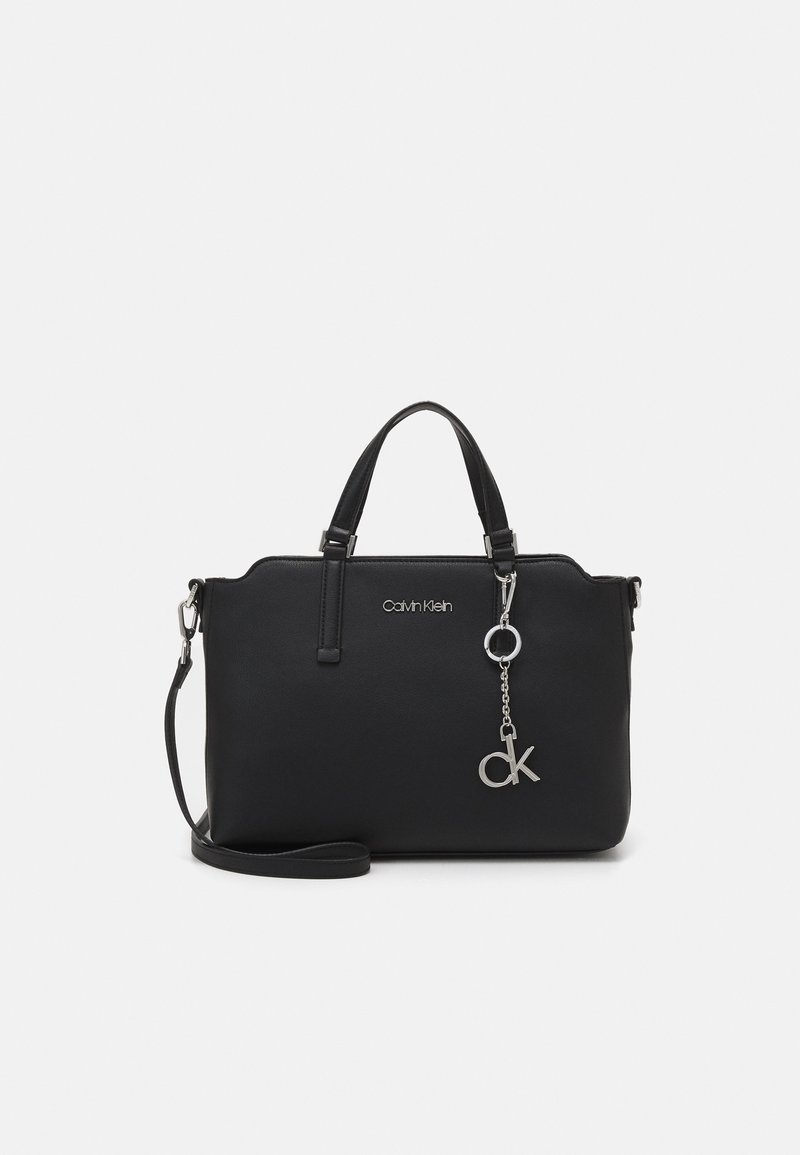 Sac à main Calvin Klein noir avec doubles poignées, bandoulière amovible et pendentif logo CK argenté sur chaîne sur fond blanc.