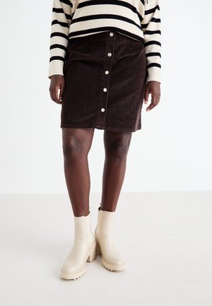Person trägt ein schwarz-weiß gestreiftes Langarmshirt, einen dunklen Cordknöpfe-Rock und cremefarbene, klobige Stiefeletten mit Absatz.