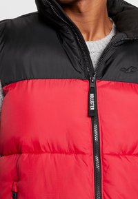 Rote und schwarze gep padded Weste mit hohem Kragen, Reißverschluss vorne und kleinem Logo auf der linken Brust. Strukturiertes Material mit gestickten Details.