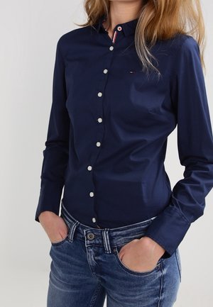 Kobieta ubrana w dopasowaną granatową koszulę na guziki z długim rękawem i niebieskie dżinsy, ręce wsunięte w przednie kieszenie.