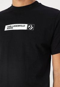 Fekete pamut póló, elöl egy fehér téglalap alakú logóval, amelyen a "KARL LAGERFELD JEANS" felirat és a "KL" grafika található. Rövid ujjak.
