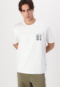 Hvit bomull t-skjorte med rund hals, korte ermer og en lomme foran. Har svart tekst som sier "GUESS JEANS 81." Enkel design, myk tekstur.
