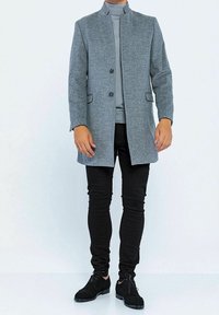 Cappotto grigio in misto lana con colletto rialzato, chiusura frontale con bottoni e tasche laterali, indossato sopra un dolcevita grigio e pantaloni neri a vestibilità slim.