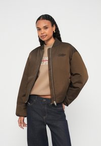 Jachetă bomber maro cu finisaj neted, guler striat și închidere cu fermoar. Asortată cu un top bej și blugi închiși la culoare. Design simplu cu broderie.