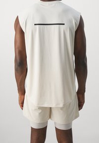 Top atletico senza maniche beige chiaro con una striscia nera orizzontale sulla schiena, abbinato a pantaloni corti beige e pantaloni intimi bianchi.