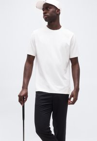 Άνδρας με λευκό καπέλο, λευκό μπλουζάκι και μαύρο παντελόνι, κρατώντας ένα γκολφ ρόπαλο, στέκεται μπροστά σε ένα απλό λευκό φόντο.