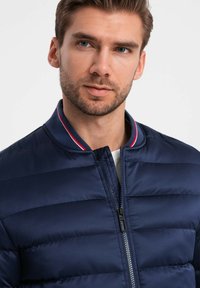 Navyblaue gepolsterte Bomberjacke mit horizontaler Steppung, einem Reißverschluss und einem kontrastierenden rot-navy gestreiften Kragen. Leicht und mit glatter Textur.