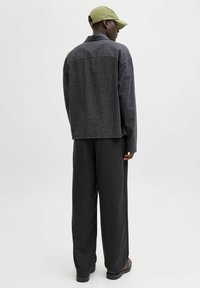 Persona con una gorra verde, chaqueta de cuadros en blanco y negro, pantalones negros holgados y zapatos marrones de pie contra un fondo blanco liso.