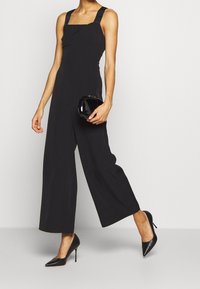 Combinaison noire avec un décolleté carré et des jambes larges, dotée de bretelles épaisses et d'une pochette noire brillante. Le modèle porte des talons noirs pointus.