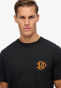 Camiseta negra de manga corta hecha de una tela suave, con un bolsillo en el pecho que presenta un logo naranja. Cuello redondo clásico y corte relajado.