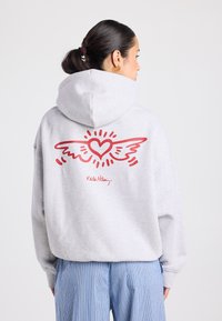 Hoodie gris avec un grand cœur rouge et un motif d'ailes au dos. Comprend une capuche à cordon de serrage et une coupe décontractée. Le texte sous le design indique "Keith Haring".