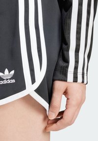 Hand som lätt håller i kanten på svarta Adidas-shorts med vita ränder och logotyp, bär en matchande svart ärm med vita ränder.