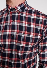 Camicia a quadri con bottoni in blu navy, rosso e bianco. Presenta un colletto classico, bottoni a contrasto e una tasca sul petto con motivo a scacchi.