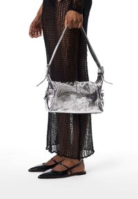 Sac à main argenté métallique à finition texturée, avec une sangle courte et des détails à fermeture éclair, tenu à côté d'une robe noire en maille et de chaussures pointues.