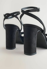 Svarta mocka klacksandaler med en kraftig klack. Har överkorsade remmar och ett spänne. Slät innersida och texturerad yttersida.