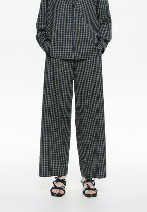 Person in weiten schwarzen Hosen und passendem karierten Hemd mit den Händen in den Taschen, kombiniert mit schwarzen und grünen Sandalen mit Riemen.