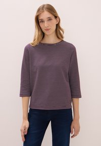 Kvinna med rakt blont hår bär en lila och vit randig tröja med trekvartsärmar och mörkblå jeans, stående mot en enkel vägg.