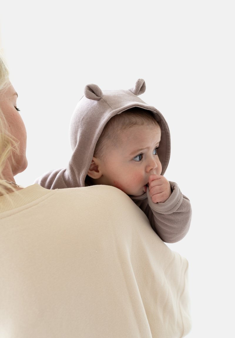 Baby trägt einen hellbraunen Kapuzenanzug mit Bärenohren und hat den Daumen im Mund. Der Hintergrund ist schneeweiß. Die Person trägt ein beiges Oberteil.