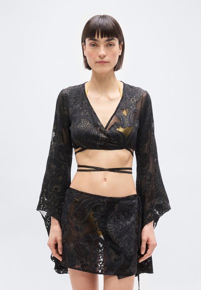Femme portant un crop top noir transparent à voile et une jupe assortie avec des motifs floraux dorés, debout devant un fond clair uni.