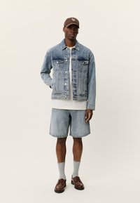 Τζιν μπουφάν σε ανοιχτό μπλε με δύο τσέπες στο στήθος, συνδυασμένο με σορτς σε ανοιχτό μπλε, γκρι κάλτσες, καφέ αθλητικά παπούτσια και καφέ καπέλο.