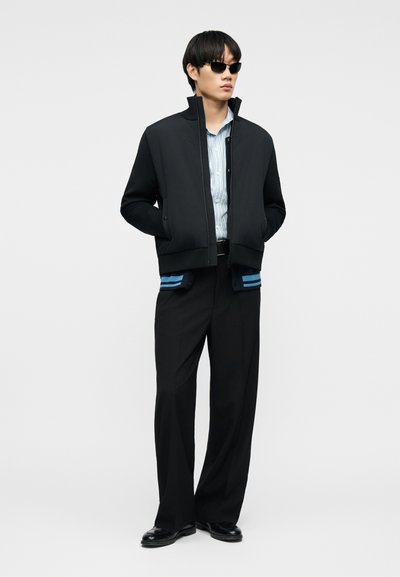 Άντρας φοράει μαύρο μπουφάν με φερμουάρ, ριγωτό μπλε πουκάμισο, μαύρο παντελόνι με φαρδιά πόδια, μαύρα παπούτσια και γυαλιά ηλίου, στέκεται με τα χέρια στις τσέπες.