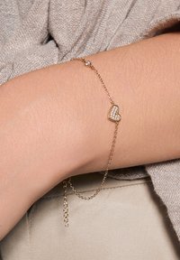 Złota bransoletka z delikatnym łańcuszkiem, ozdobiona wisiorkiem w kształcie serca, przyozdobionym małymi, przezroczystymi kamieniami szlachetnymi. Gładka tekstura i minimalistyczny design.