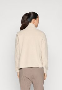 Chaqueta de forro polar beige con cuello alzado, tejido texturizado y un corte cropped, combinada con pantalones marrones claros. Detalle de costura sutil.