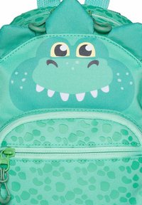 Mochila temática de dinosaurio verde con una cara sonriente, tela texturizada, una parte superior con picos, un compartimento principal y un pequeño bolsillo frontal con cremallera.