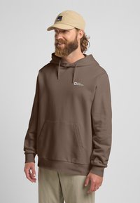 Felpa marrone con tasca frontale, cappuccio regolabile e logo. Abbinata a un cappellino beige, che presenta un look casual e texturizzato.