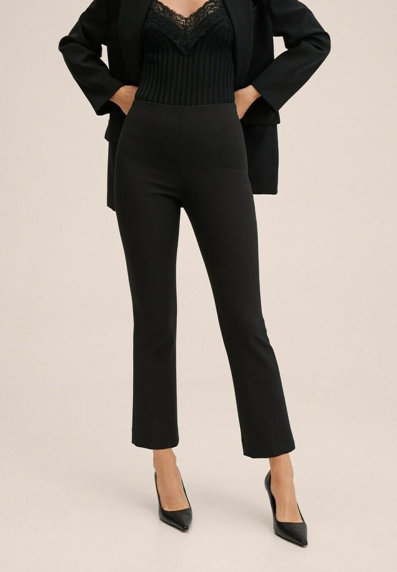 Femme portant un haut noir ajusté en maille côtelée orné de dentelle, un pantalon court noir, un blazer noir et des escarpins noirs à bouts pointus, mains dans les poches.