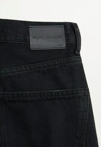 Schwarze Jeans aus Denim mit einem ledernen Etikett, das "BLUE EDITION" trägt. Auffällige Nähte und Taschen sind sichtbar und unterstreichen ein klares Design.