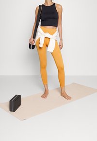Mujer de pie descalza sobre una esterilla de yoga beige, llevando un top corto negro, leggings naranjas, un suéter blanco atado a la cintura y cargando una bolsa negra.