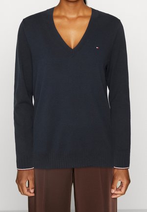 Femme portant un pull col V bleu marine avec un petit logo rouge, blanc et bleu sur la poitrine et un pantalon marron, debout devant un fond uni.