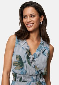 Ärmellose Bluse mit Floral-Muster und Rüschenausschnitt, die grüne und blaue Blätter auf einem gestreiften, hellen Hintergrund zeigt und einen elastischen Bund hat.