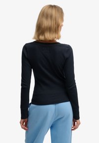 Haut noir à manches longues avec un design simple, fabriqué dans un tissu lisse. Le dos comporte une petite étiquette carrée au-dessus de l'encolure.