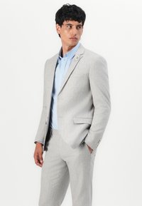 Ανοιχτό γκρι κοστούμι με κομψή εφαρμογή, που διαθέτει σακάκι με ένα κουμπί, γιακά notch και ανοιχτό μπλε πουκάμισο από κάτω.