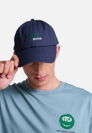 Młoda osoba nosząca granatową czapkę z napisem "THANKS 4 NOTHING CLUB RVCA" oraz jasnoniebieską koszulę z zielonym logo RVCA i napisem.
