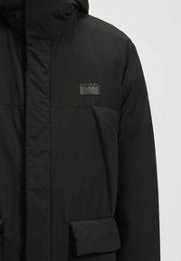 Giacca imbottita nera con cappuccio, dotata di un patch logo nero opaco, tasche frontali e un tessuto strutturato per isolamento.