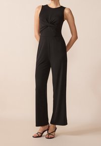 Mono negro con un torso ajustado y detalle de torsión en el frente; diseño de pantalón de pierna ancha; tela suave y elástica; combinado con sandalias negras de tiras.