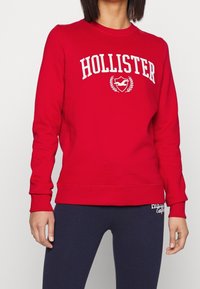 Sudadera roja con el logo blanco de "HOLLISTER", escote redondo y puños acanalados. Combinada con leggings azul marino que tienen acentos blancos.