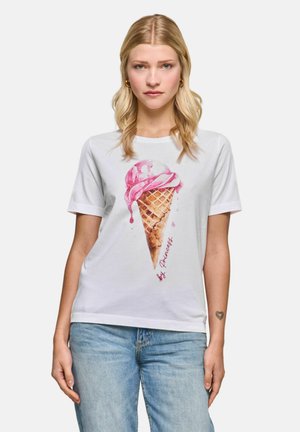 Woman with blond hair wears a white t-shirt with a large pink ice cream cone graphic and blue jeans, standing against a plain background.