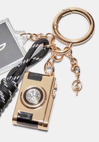 Portachiavi con una miniatura di una macchina fotografica dorata con dettagli neri, decorata con una pietra trasparente. Include un cinturino intrecciato in bianco e nero.