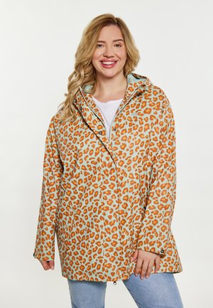Nainen hymyilee, pukeutunut oranssi-beige leopardikuvioiseen hupulliseen sadetakkiin vaaleansinisten farkkujen ja valkoisen paidan päällä.